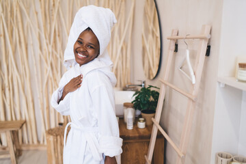 Half length portrait of joyful black woman in cozy bathrobe enjoying skincare routine at home during authentic moment in contemporary bathroom with natural decor elements, expression emotions