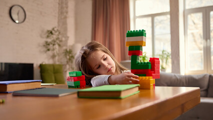 Child girl sitting at desk at home in living room, playing with building blocks and notebooks. Focus and concentration during play. Concept pf childhood, early development, learning activity