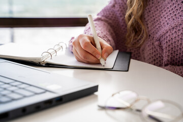 Female hand writing with pen in notebook on desk with laptop and eyeglasses, unrecognizable...