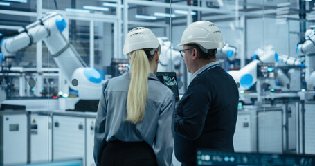 Obraz premium Electronics Factory: Female Project Manager and Engineer in Hard Hats, Talking, Using Laptop. Monitoring Advanced Equipment Production. Robotic Arms Assembly Line Manufacturing Circuit Boards