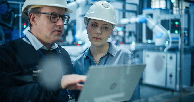 System Administrator and Technician Working at a Manufacturing Plant. Male Specialist Programming the Work of the Robotic Arms, Explaining Production on the Assembly Conveyor to a Female Colleague - Powered by Adobe