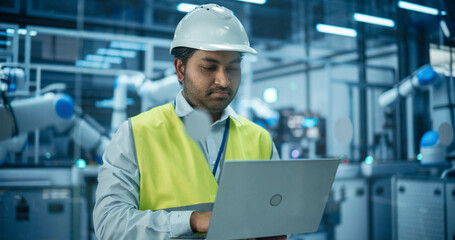 Fototapeta premium Portrait of an Industrial Systems Administrator Working on Laptop Computer at a Modern Manufacturing Plant. Indian Specialist in a Hard Hat and High Visibility Vest Optimizing Manufacturing Flow