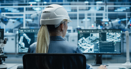 Female Engineer Sitting Behind a Desk in a Modern Product Development Facility, Using Industrial Computer Software on Dual Screens, Monitoring Electronics Production Line with Robotic Arms
