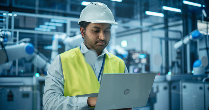 Portrait of an Industrial Systems Administrator Working on Laptop Computer at a Modern Manufacturing Plant. Indian Specialist in a Hard Hat and High Visibility Vest Optimizing Manufacturing Flow - Powered by Adobe