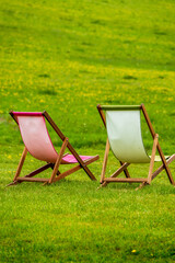Two wooden deck chairs with pastel canvas slings arranged on a verdant grassy slope beneath...