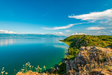 Emerald shoreline curves under limestone promontory at Lin, Albania on tranquil Lake Ohrid beneath an expansive cerulean sky
