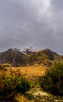 Snow-dusted Glyderau ridge rising above a heather-clad corrie basin and lichen-scarred boulders under a brooding Welsh sky