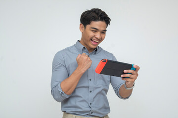 A happy young Southeast Asian man in smart casual wear pumped at winning while playing on a handheld gaming console. Isolated on a light background. © Mdv Edwards