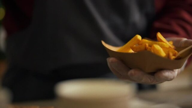 A person in gloves carefully portions crispy fries into a takeout container at a vibrant food stall. The busy atmosphere of the festival creates excitement as attendees eagerly wait for tasty treats