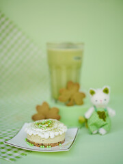 Matcha menu. Matcha greentea cheesecake mini tart on the foreground and Matcha green tea latte on the background. Soft focus on the cheesecake. Shamrock cookies decorations for Saint Patrick's day