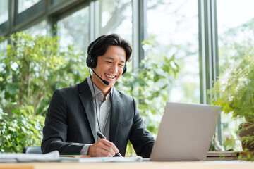 Asian businessman wearing a headset, smiling while talking to a laptop, holding a pen, bright office with large window, fresh greenery outside, modern remote work atmosphere no logo