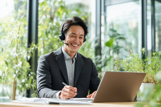 Asian businessman wearing a headset, smiling while talking to a laptop, holding a pen, bright office with large window, fresh greenery outside, modern remote work atmosphere no logo