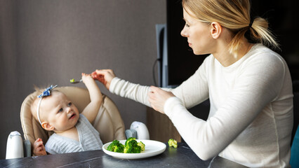 Mother trying to feed baby with spoon healthy broccoli at home, toddler resisting and showing displeasure. Authentic parenting lifestyle, indoor struggle with nutrition. Real family moment with child