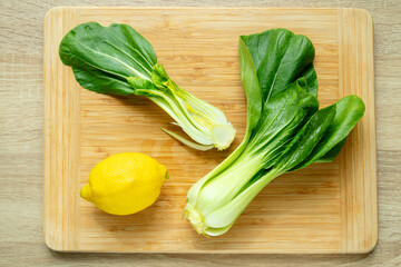 Cut green pak choi cabbage leaves and fresh lemon on wooden board. Fresh Chinese cabbage pak choi in the process of cooking. Top view.