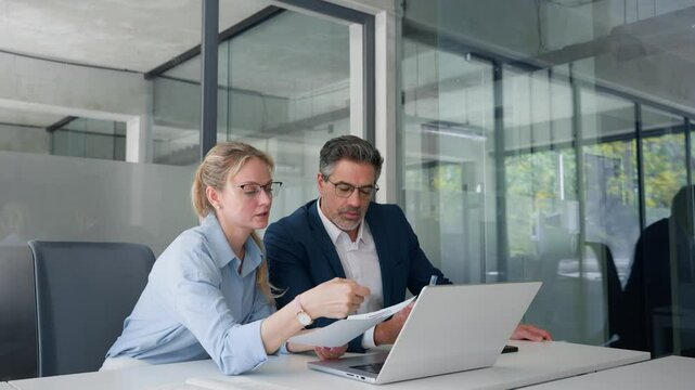 Team of diverse partners mature man and young woman discussing financial document using computer sitting at table in office. Two colleagues of professional business people working together on project