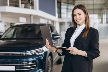 Saleswoman holding laptop is posing in car dealership