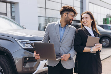 Car salespeople working together holding laptop and tablet in front of new car