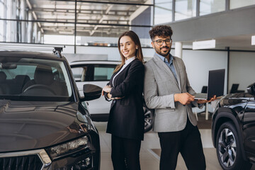 Car salespeople standing back to back in a dealership, holding a laptop