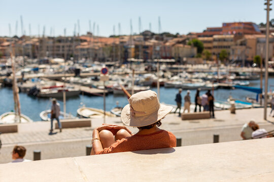 Femme en vacances portant un chapeau assise tranquillement sur des marches au soleil regardant la ville en premier plan, avec le port et des bateaux en arri&egrave;re plan
