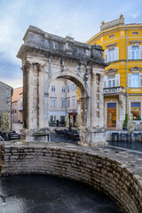 The historic Roman Gate in Pula, Croatia, with colorful buildings lining the square and outdoor seating nearby.