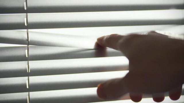 A hand is seen carefully gripping the edge of window blinds, pulling them aside to allow sunlight to illuminate the room. This moment captures a peaceful morning in a domestic space