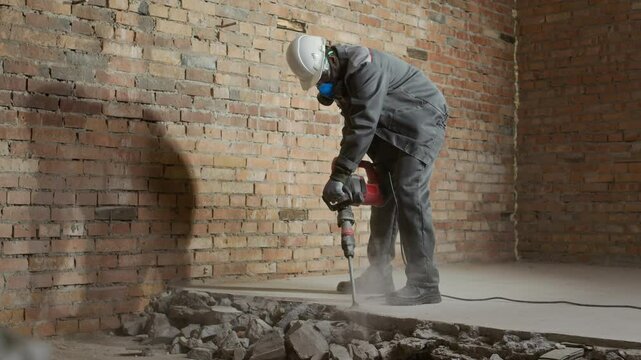 Low angle view of male construction worker wearing hardhat and respirator destroying floors with demolition hammer inside unfinished room