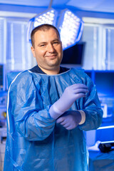 Med professional readying for procedure. A man in a blue medical gown puts on gloves in a modern...