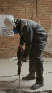 Vertical shot of male construction worker in goggles, hardhat and respirator using jackhammer while destroying floor on worksite
