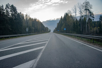 Naklejka premium An empty highway surrounded by forest stretches into the distance under a dramatic cloudy sky, with white road markings and metal guardrails.