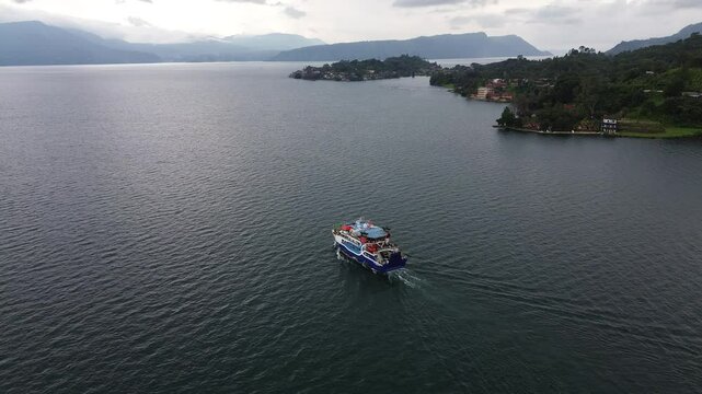 Aerial view of ferry crossing Lake Toba from Parapat to Ambarita, Samosir Island, North Sumatra, Indonesia, with scenic cloudy sky and calm water reflection