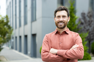 A smiling man in an orange shirt stands in front of a modern building, looking confident with arms...