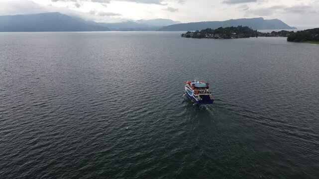 Aerial view of ferry crossing Lake Toba from Parapat to Ambarita, Samosir Island, North Sumatra, Indonesia, with scenic cloudy sky and calm water reflection