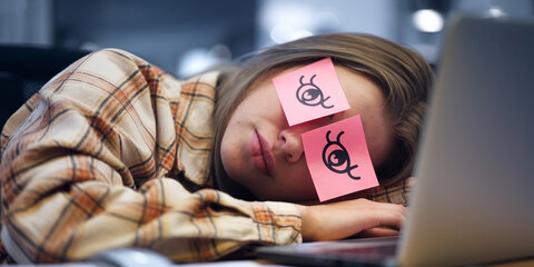 Young woman sleeping on desk, pink sticky notes with eye drawings over her eyelids, wearing plaid shirt, laptop nearby, showcasing exhaustion, stress, or need for break