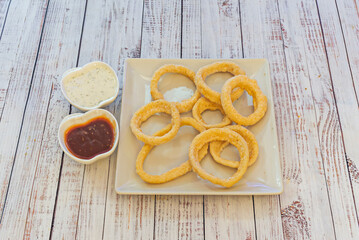 A square plate filled with fried onion rings along with a couple of dipping sauces