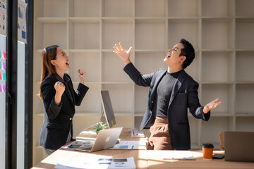Two people are standing in front of a desk with a laptop and a cup
