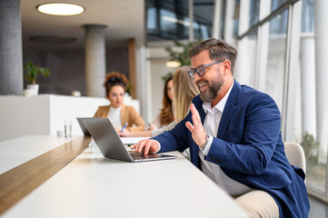Businessman in glasses waving hand during video call on his laptop while female colleagues working in the background