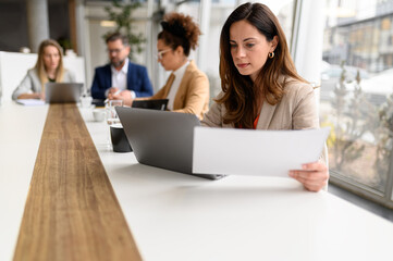 Focused accountant holding report and working over laptop while teammates collaborating in the background