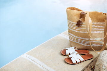 Vacation Concept. Straw beach bag with white sandals placed neatly by swimming pool, the calm blue...