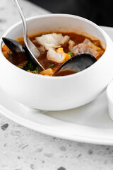 A close-up of a seafood soup in a white bowl, featuring fish, mussels, and a rich red broth, garnished with fresh parsley. The dish is accompanied by a spoon and a mussel shell.