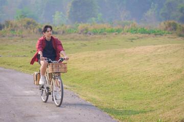Naklejka premium A Handsome young man in a red shirt and black sports shorts is happily riding a white bicycle in the morning for exercise and relaxing in the park during holiday. Healthy exercise concept.