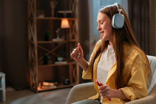 Young teenager girl spending evening time at home, girl sits in chair in the living room and listening music in headphones, dancing positive expression.