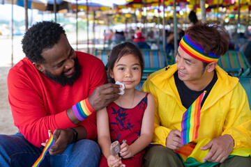 african and asian male gay couple sitting on beach chair are taking care female kid daughter eating ice cream while travel on summer trip