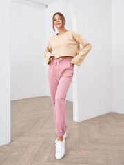 A young woman stands confidently in a minimalistic setting, wearing a beige crop top, pink joggers,...
