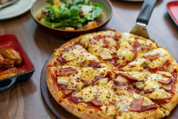 scooping hawaiian pizza dough thick and soft with cheese pineapple ham on wooden tray and grilled chicken drumstick barbecue with vegetable salad for lunch to family eating food at italian restaurant