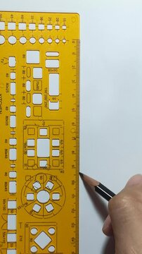 A hand holds a pencil next to a yellow template ruler on white paper, ready to draw or measure precisely.