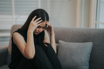 Depressed and unhappy Asian woman sits with anxiety on the sofa in her living room at home.