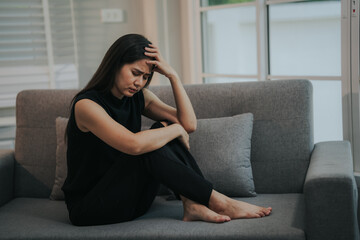 Obraz premium Depressed and unhappy Asian woman sits with anxiety on the sofa in her living room at home.