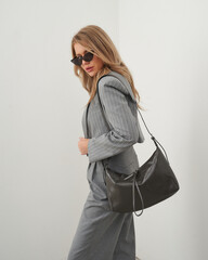 Posing blonde woman in stylish blazer and trousers with handbag in modern studio setting