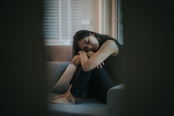 Fototapeta premium Depressed and unhappy Asian woman sits with anxiety on the sofa in her living room at home.