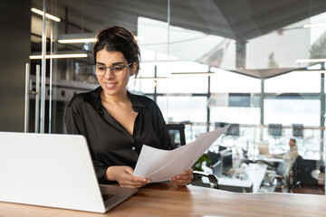 Smiling latin or arabian young business woman hr working on laptop computer reading finance document report, cv contract in office. Accountant saleswoman manager businesswoman doing paperwork using pc